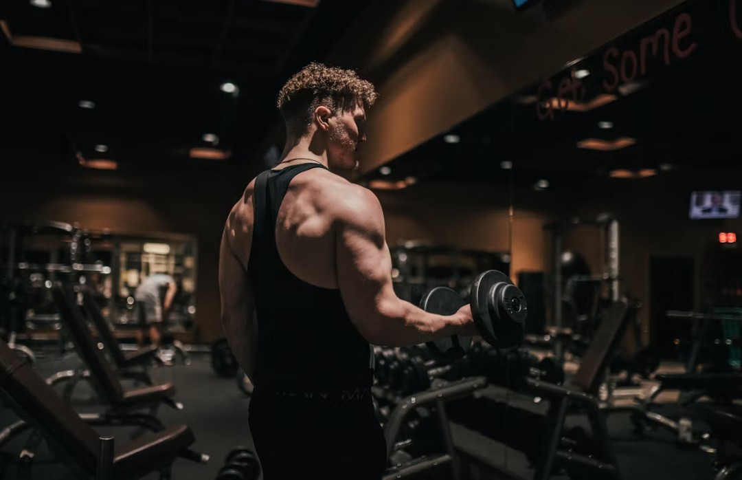 Hombre musculoso de espaldas haciendo curl de bíceps con una mancuerna en un gimnasio